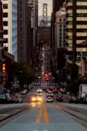A steep downtown street with cable car tracks at dusk, the Bay Bridge at its far end